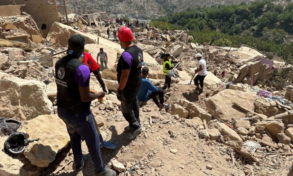 Des membres de l'équipe Ummah Charity se tiennent sur des maisons détruites, apportant leur aide et leur soutien à la population touchée par le séisme au Maroc, illustrant l'engagement humanitaire en période de crise.